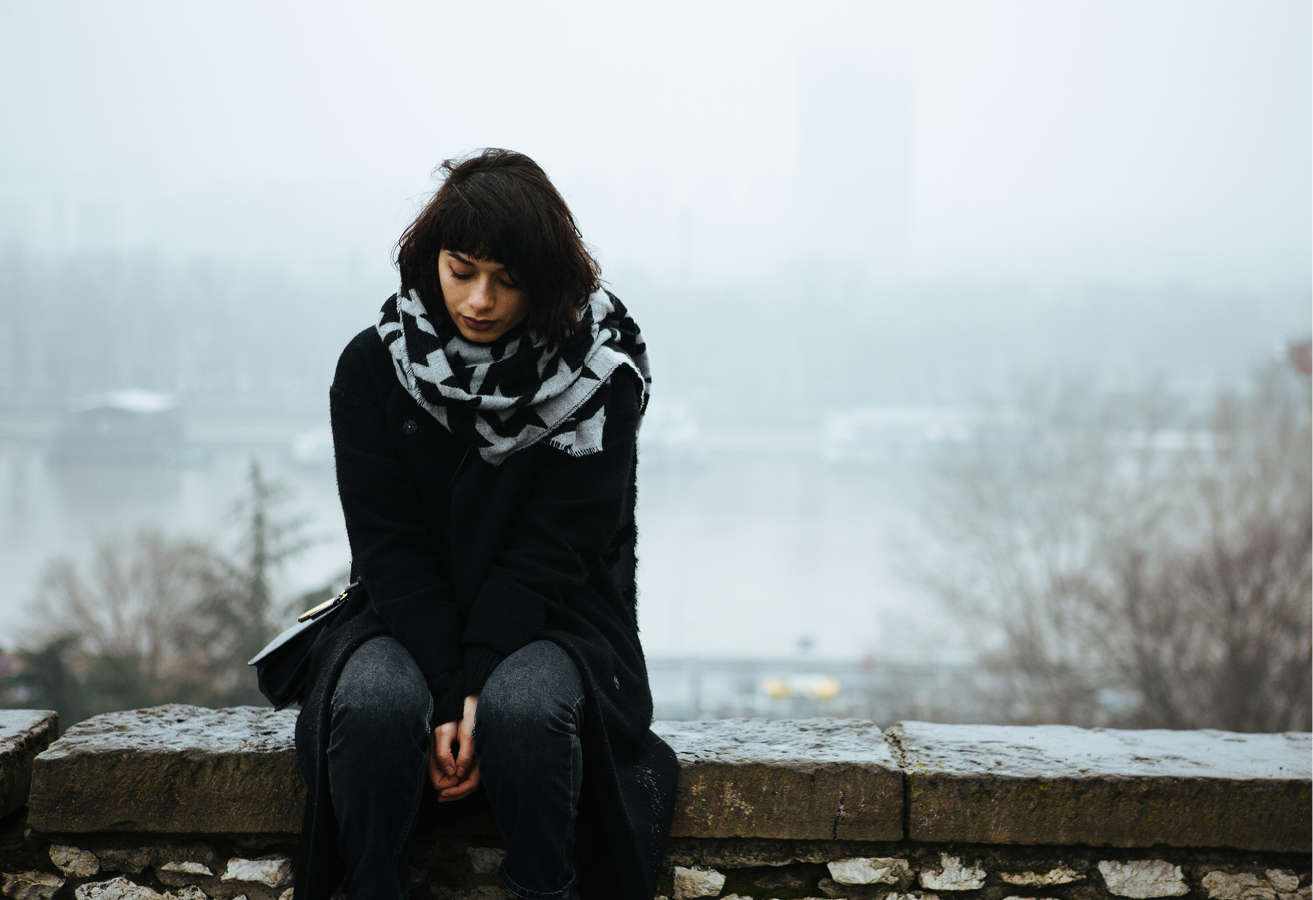 Woman sitting alone on a stone wall, wearing dark clothes, looking down and appearing sad. The background is gloomy weather.